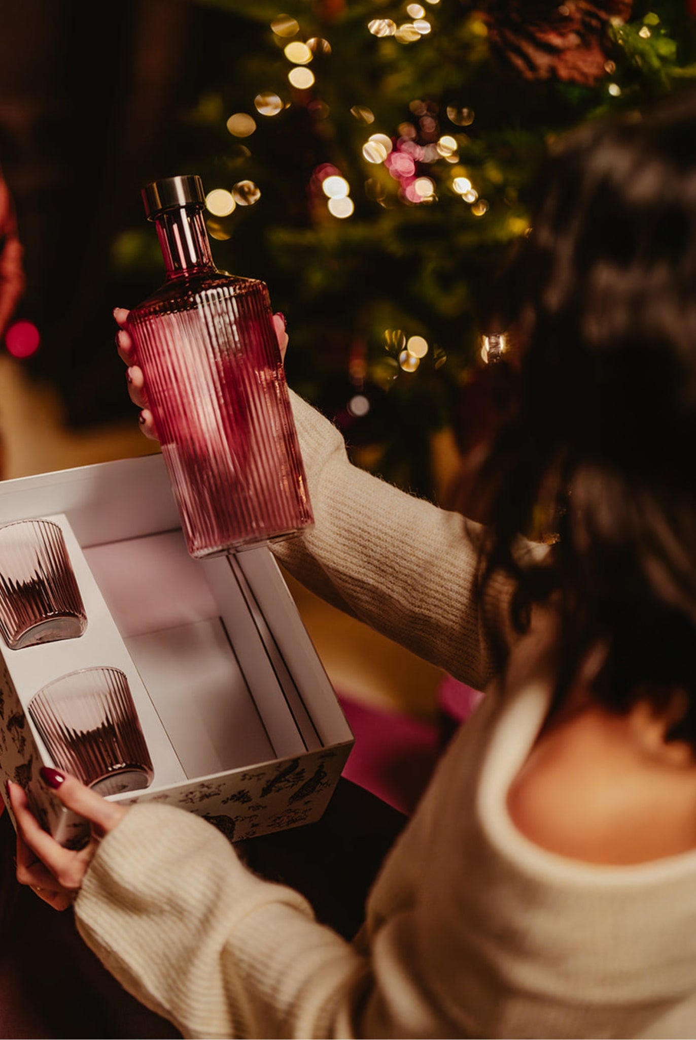 Eine Dame öffnet das Geschenkset Paveau und hält die Paveau Glasflasche in Amethyst in ihrer Hand, neben einem Weihnachtsbaum.