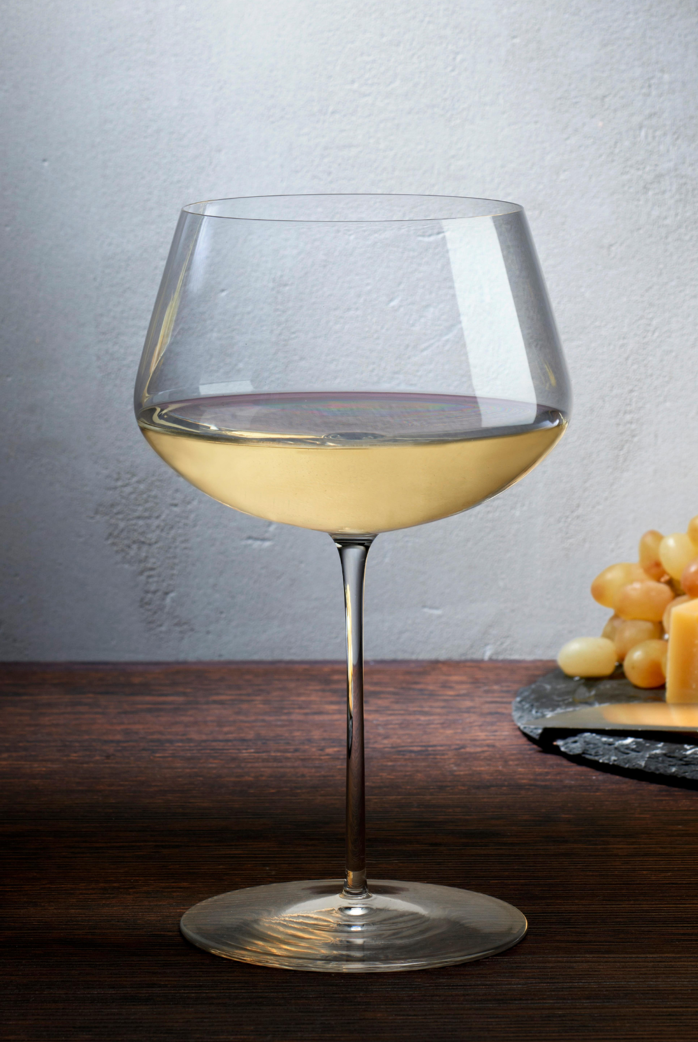 Weißweinglas transparent mit Weißwein gefüllt auf Tisch stehend neben Teller mit Käse und Weintrauben