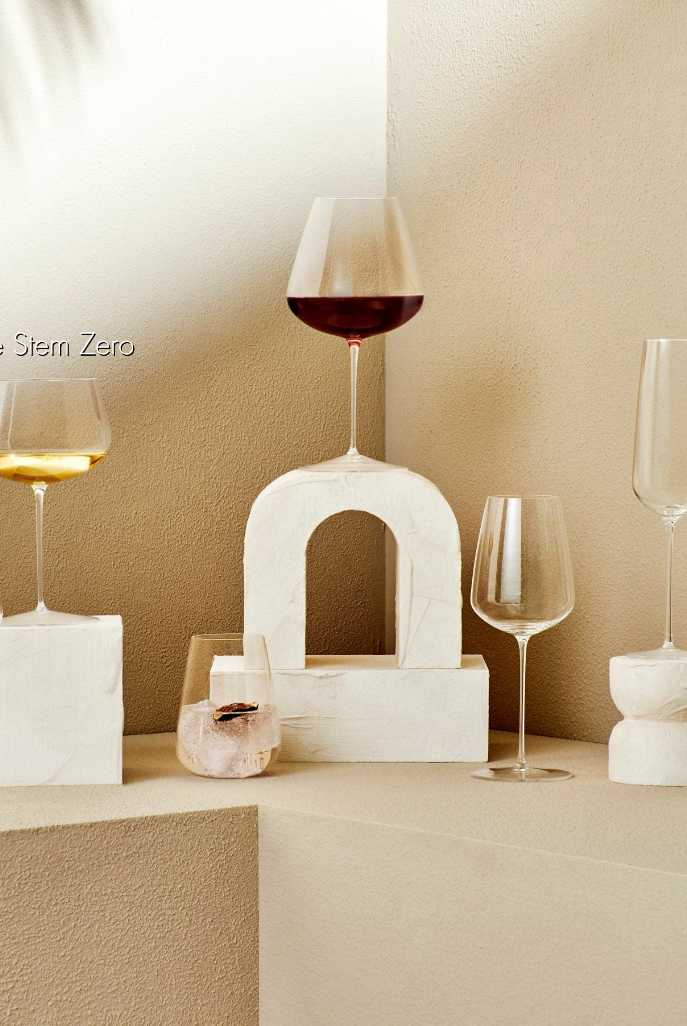 Weißweinglas transparent mit Weißwein gefüllt auf steinernem Viereck weiß stehend neben sechs transparenten Champagnergläsern, Weißweingläsern, Rotweingläsern und Wassergläsern mit sommerlichen Hintergrund