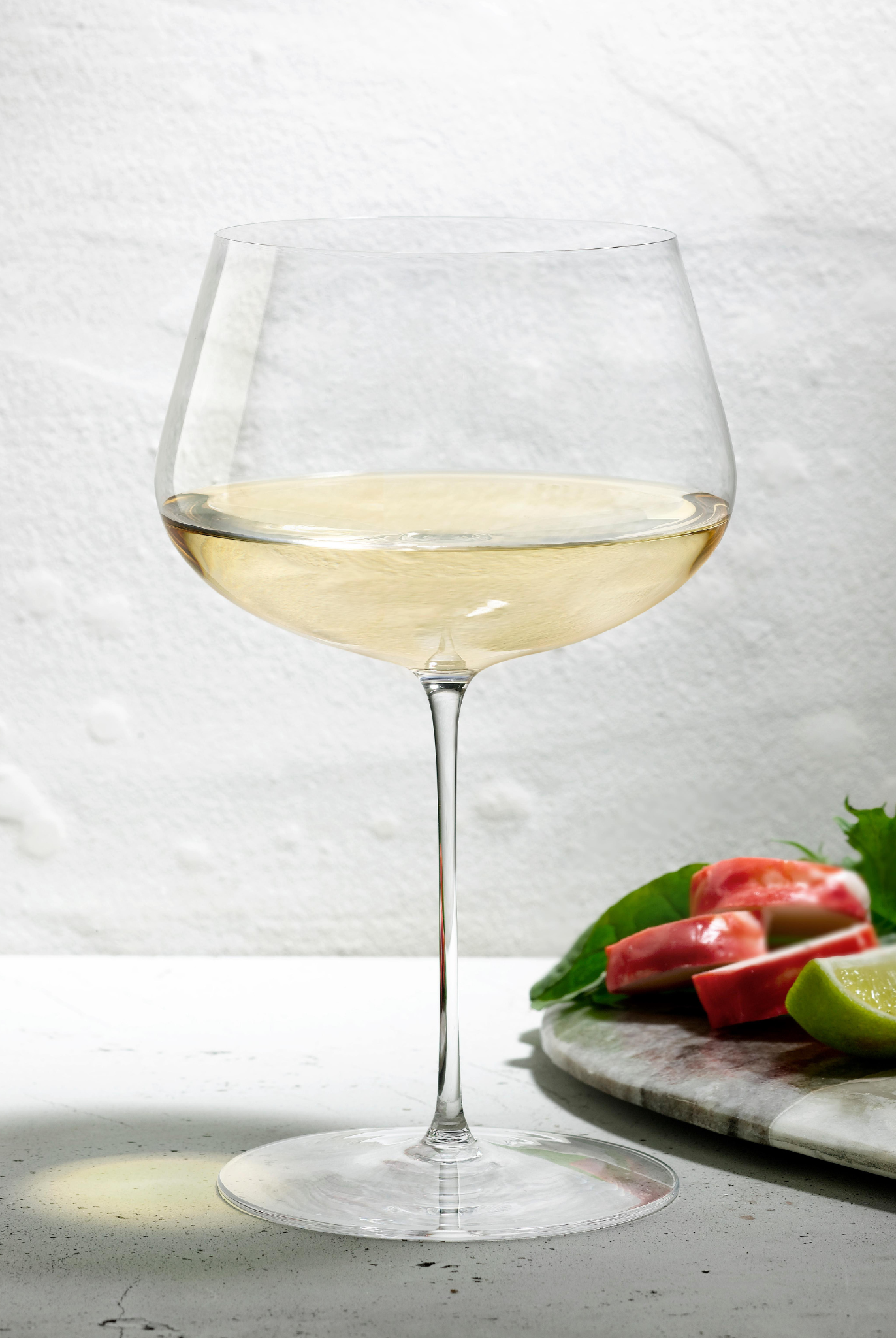 Weißweinglas transparent mit Weißwein gefüllt auf Tisch stehend neben Brett mit geschnittenen Früchten