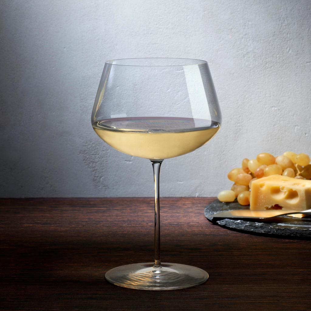 Weißweinglas transparent mit Weißwein gefüllt auf Tisch stehend neben Teller mit Käse und Weintrauben
