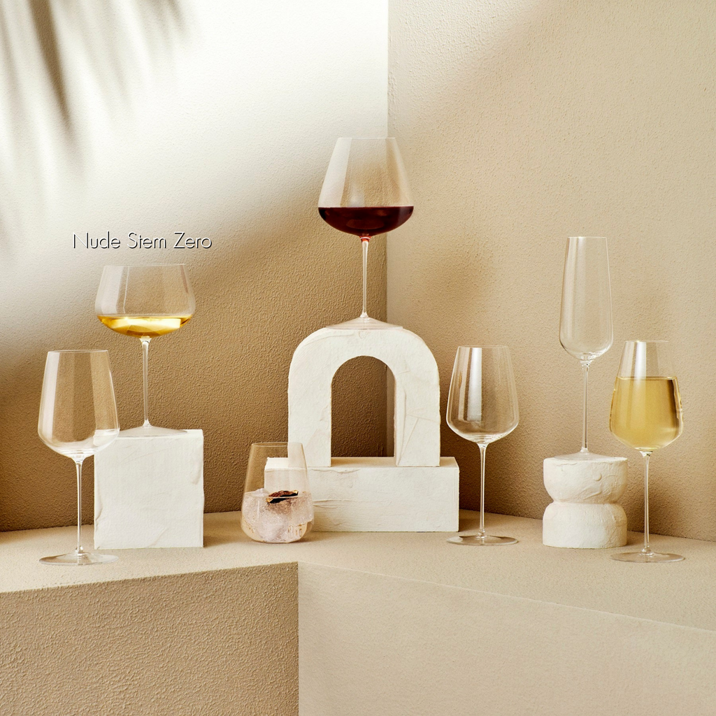 Weißweinglas transparent mit Weißwein gefüllt auf steinernem Viereck weiß stehend neben sechs transparenten Champagnergläsern, Weißweingläsern, Rotweingläsern und Wassergläsern mit sommerlichen Hintergrund